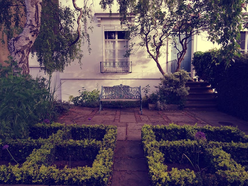 A lush, small front garden featuring a variety of greenery, including large-leaved plants, dense shrubs, and mature trees providing shade. There is a neatly trimmed lawn with a small paved seating are