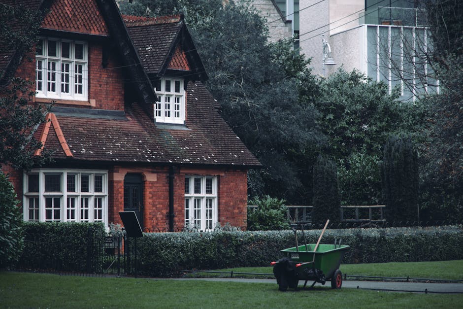 A red brick, two-story house with a steep, tiled roof and white-framed windows is situated in a well-maintained garden. The garden features a neatly trimmed hedge bordering a grassy lawn, with a pathw